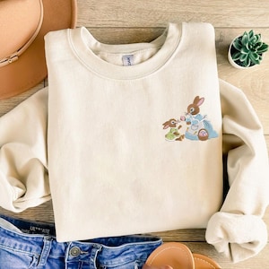 Könnte beinhalten: Cremefarbenes Sweatshirt mit einem gestickten Osterhasen-Design. Das Design zeigt zwei Hasen, einen in einem blauen Kleid und den anderen in einem grünen Kleid, mit einem Osterkorb. Das Sweatshirt ist ein lässiges Oberteil mit langen Ärmeln.