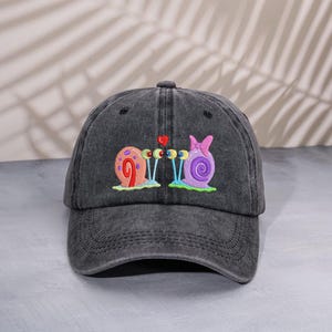 Può includere: Cappellino da baseball grigio antracite con un design ricamato di due lumache con antenne a forma di cuore. Le lumache sono in tonalità rosa, viola e arancione, con occhi blu e dettagli verdi. Il cappellino ha una visiera curva e un cinturino regolabile.