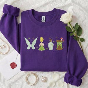 Könnte beinhalten: Ein lila Sweatshirt mit gestickten Designs zum Thema Feen. Das Design umfasst Feenflügel, eine Fee, eine Zaubertrankflasche und ein Pilzhaus. Das Sweatshirt hat gerollte Ärmel und einen Rundhalsausschnitt.