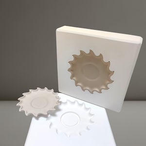 Op de afbeelding: Een set witte keramische mallen en een afgewerkte plaat. De mallen zijn vierkant met een uitgesneden sunburst-ontwerp en de plaat heeft een bijpassende sunburst-rand. De plaat is gebroken wit en de mallen zijn helderwit.