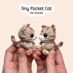 May include: Two small crocheted tabby cats with beige and brown yarn, black plastic eyes, and pink stitched paws. The text Tiny Pocket Cat PDF PATTERN is displayed above the figures. The cats have pointed ears, striped markings, and tails.