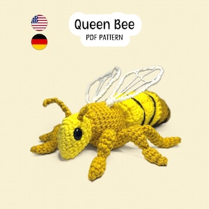 Puede incluir: Un juguete de abeja tejido a ganchillo con cabeza, tórax y abdomen amarillos con rayas negras. Tiene alas de hilo blanco y ojos de cuentas negras. El texto superior dice Queen Bee PDF PATTERN, junto a iconos de banderas estadounidense y alemana.