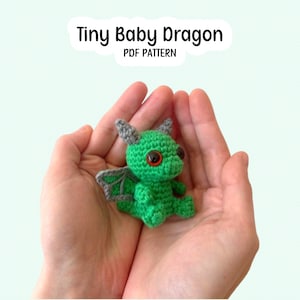 May include: A small green crocheted dragon toy with grey wings and large orange eyes. The text Tiny Baby Dragon PDF PATTERN is displayed above the toy. The dragon has a textured yarn surface and is crafted with detailed stitching throughout.