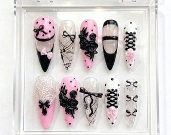 Peut inclure: Un ensemble de dix faux ongles dans un étui en plastique transparent. Les ongles présentent divers motifs, notamment des nœuds noirs, de la dentelle et des roses, avec une palette de couleurs rose, noir et blanc. Certains ongles ont une pointe noire.
