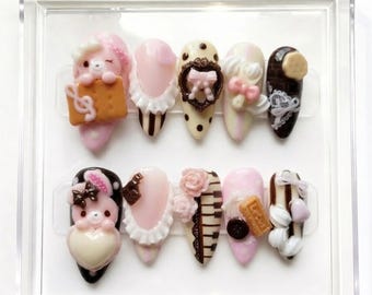 May include: A set of ten artificial nails in a clear plastic case. The nails are decorated with various designs, including a bear, bows, hearts, and musical notes, in shades of pink, white, and brown.