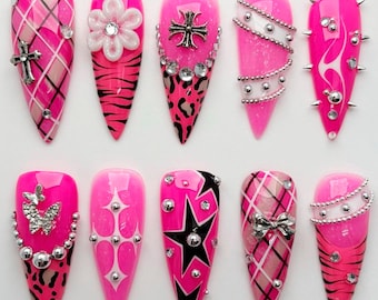 May include: A set of ten long, pointed, artificial nails in vibrant pink. Each nail features unique designs, including crosses, stars, bows, and animal prints, adorned with rhinestones and silver accents. Some nails have a plaid pattern or a flame design.