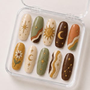 Puede incluir: Un juego de diez uñas postizas ovaladas dispuestas en un estuche de plástico transparente. Los diseños presentan motivos celestiales que incluyen soles, lunas y estrellas, junto con patrones florales y líneas onduladas en crema, verde salvia y marrón.