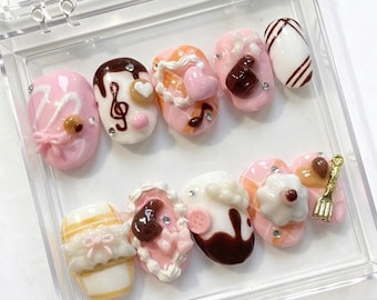 May include: A set of ten artificial nails in a clear plastic case. The nails are decorated with various dessert-themed designs, including pink frosting, chocolate drizzle, musical notes, and heart shapes. Some nails feature rhinestones and miniature decorative elements.