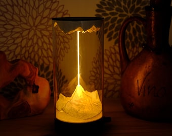 May include: A cylindrical lamp with a mountain-like design at the base, illuminated from within. The lamp is made of clear material, with a black base and top. A decorative jug and sculpture are in the background.