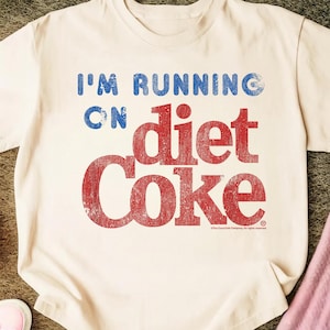 May include: Cream-colored t-shirt with the text "I'M RUNNING ON diet Coke" in blue and red distressed lettering. The shirt has a classic crew neck and short sleeves. The Coca-Cola Company logo is at the bottom right.