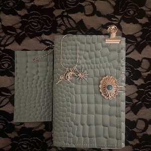 Puede incluir: Un cuaderno azul claro con textura de cocodrilo y un pequeño tarjetero a juego. El cuaderno cuenta con un clip de metal plateado, un dije de sol y luna en un lazo de alambre y un concho ovalado plateado con una piedra turquesa central.