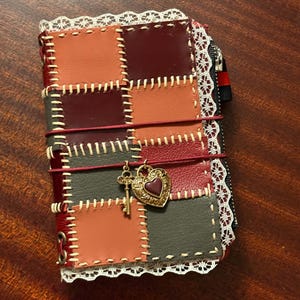 May include: A patchwork journal in red, orange, and grey leather. It features a gold heart charm with a key pendant, and is edged with white lace. A red elastic band holds the journal closed. The journal is a unique handmade item.