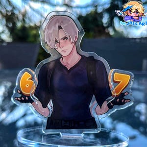 Puede incluir: Un expositor de acrílico transparente con un personaje de estilo anime de pelo claro y camiseta oscura, que sostiene los números naranjas 6 y 7 en cada mano. El personaje tiene las mejillas sonrojadas y lleva guantes negros sin dedos.