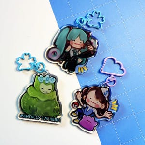 May include: Three acrylic keychains featuring cartoon characters. One keychain depicts a green character with the text "MENTALLY I'M HERE". Another shows a girl with a pink Ditto. The third keychain shows a girl with teal pigtails holding a microphone.