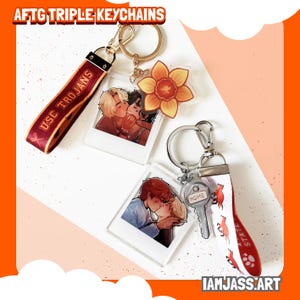 May include: AFTG triple keychains with various designs. One keychain features a maroon strap with "USC Trojans" text. Other keychains include illustrations of couples kissing, a daffodil, and a key with "HOME" text. The artist's name, IAMJASS.ART, is visible.