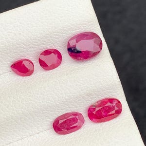 May include: An assortment of polished, red ruby gemstones of varying sizes and shapes. The gemstones are displayed on a white surface against a dark background. The rubies include oval and round cuts.