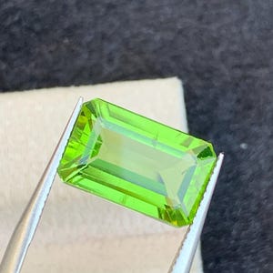 Puede incluir: Una gema rectangular verde brillante sostenida por pinzas plateadas. La gema tiene un corte facetado, que refleja la luz. El fondo es una superficie oscura y texturizada con un paño blanco.