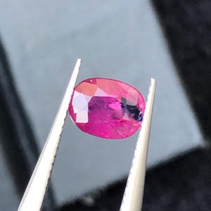 May include: A vibrant, oval-cut gemstone with a deep magenta and pink hue, featuring multiple facets that reflect light. The stone is held securely by a pair of silver-toned metal tweezers, showcasing its translucent quality and polished surface.