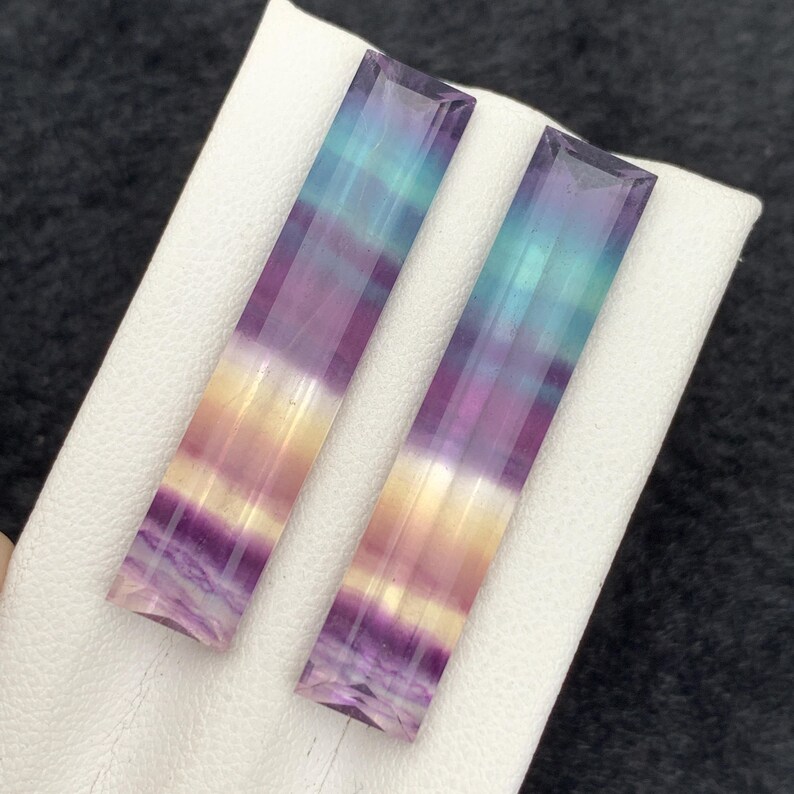 K&ouml;nnte beinhalten: Zwei rechteckige Fluorite mit Farbverlauf. Die Steine zeigen B&auml;nder in Lila, Gelb, Blau und Gr&uuml;n. Sie liegen auf einer wei&szlig;en Oberfl&auml;che vor dunklem Hintergrund.