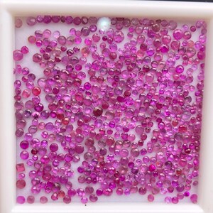 May include: A collection of numerous small, round, faceted pink and magenta gemstones arranged in a white square display tray. The stones vary slightly in size and depth of color, featuring a brilliant cut that reflects light across the surface.