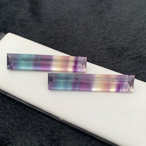 K&ouml;nnte beinhalten: Zwei rechteckige Fluorite-Edelsteine. Die Steine zeigen B&auml;nder in Lila, Gr&uuml;n und Gelb. Die Edelsteine liegen auf einer wei&szlig;en Oberfl&auml;che mit dunklem Hintergrund.