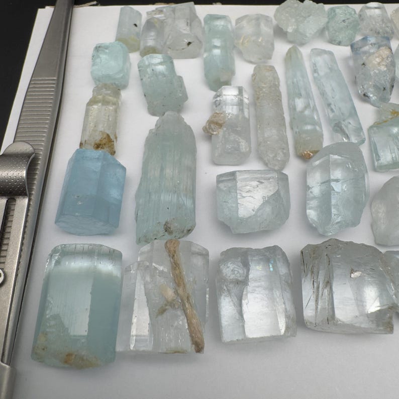 Puede incluir: Una colecci&oacute;n de cristales de aguamarina en bruto de diversas formas y tama&ntilde;os. Los cristales var&iacute;an en color de azul p&aacute;lido a transparente, algunos con inclusiones naturales. Se ve un par de pinzas met&aacute;licas en el lado izquierdo.