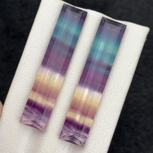 K&ouml;nnte beinhalten: Zwei rechteckige Fluorite mit Farbverlauf. Die Steine gehen von Lila &uuml;ber Gelb zu T&uuml;rkis &uuml;ber. Die Edelsteine werden auf einer wei&szlig;en Oberfl&auml;che pr&auml;sentiert.