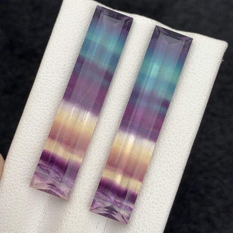 K&ouml;nnte beinhalten: Zwei rechteckige Fluorite mit Farbverlauf. Die Steine gehen von Lila &uuml;ber Gelb zu T&uuml;rkis &uuml;ber. Die Edelsteine werden auf einer wei&szlig;en Oberfl&auml;che pr&auml;sentiert.