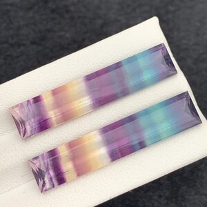 K&ouml;nnte beinhalten: Zwei rechteckige Fluorite mit einem Farbverlauf aus Lila, Gelb und Blau. Die Edelsteine sind facettiert und haben ein poliertes Aussehen. Sie werden auf einer wei&szlig;en Oberfl&auml;che pr&auml;sentiert.
