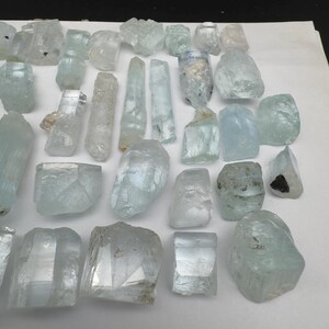 Puede incluir: Una colecci&oacute;n de cristales de aguamarina en bruto de diversas formas y tama&ntilde;os. Los cristales var&iacute;an en color, desde transparente hasta azul verdoso p&aacute;lido, con texturas y facetas naturales visibles. Algunos cristales son alargados, otros m&aacute;s redondeados.