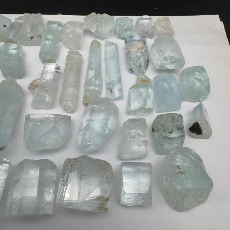 Puede incluir: Una colecci&oacute;n de cristales de aguamarina en bruto de diversas formas y tama&ntilde;os. Los cristales var&iacute;an en color, desde transparente hasta azul verdoso p&aacute;lido, con texturas y facetas naturales visibles. Algunos cristales son alargados, otros m&aacute;s redondeados.