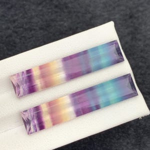 K&ouml;nnte beinhalten: Zwei rechteckige Fluorite mit einem Farbverlauf aus Lila, Blau, Gelb und Klar. Die Edelsteine werden auf einer wei&szlig;en Oberfl&auml;che mit dunklem Hintergrund pr&auml;sentiert. Die Steine haben einen facettierten Schliff.
