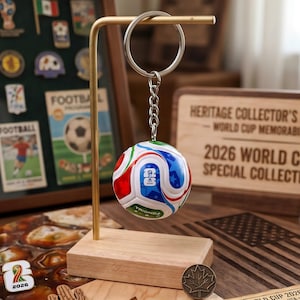 Puede incluir: Un llavero con forma de balón de fútbol con detalles en rojo, blanco, azul y verde, colgado de un soporte dorado. El llavero tiene el texto "TRIONGA" y un logotipo. El fondo incluye un letrero de madera que dice "2026 WORLD CUP SPECIAL COLLECTION."