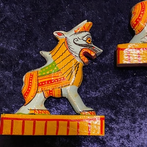 May include: Two hand-painted wooden figurines of mythical creatures with yellow, red, green, and blue details. The figurines are standing on yellow and red bases.