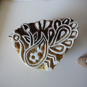 May include: A wooden block printing stamp with a detailed design of a peacock with its tail feathers spread out. The stamp is brown and white and has a textured surface.