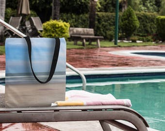 Puede incluir: Una bolsa de mano negra con una escena de playa impresa en ella. La bolsa está sentada en una silla de salón junto a una piscina.