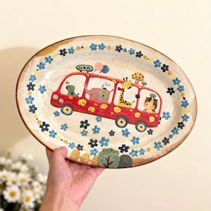 Puede incluir: Plato ovalado de cerámica con un diseño caprichoso. Un autobús rojo transporta animales de dibujos animados, incluyendo una jirafa y un león. El borde presenta patrones florales y de estrellas azules y amarillos. El plato es sostenido por una mano.