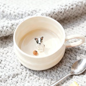 May include: Cream-colored ceramic mug with a small, detailed figurine of a dog and a miniature basketball inside. The dog is black, white, and tan. The mug has a curved handle and sits on a gray knitted surface.