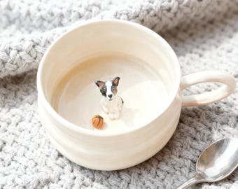 Puede incluir: Taza de cerámica color crema con una pequeña figura detallada de un perro y una pelota de baloncesto en miniatura en su interior. El perro es negro, blanco y marrón. La taza tiene un asa curvada.