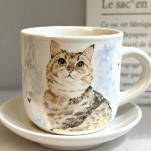 May include: A white ceramic mug and saucer set. The mug features a detailed illustration of a tabby cat with green eyes, set against a soft blue and white background. Small red hearts are scattered around the mug. The saucer is plain white.