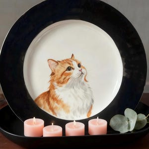 Op de afbeelding: Een decoratieve plaat met een zwarte rand en een witte binnenkant met een illustratie van een oranje en witte kat. De plaat staat op een zwart dienblad met vier roze kaarsen en een takje groen.
