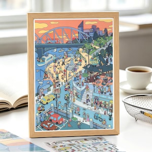 Op de afbeelding: Een ingelijste illustratie van een drukke stadsscene aan het water met een grote brug, een bistro, diverse voetgangers, auto's en fietsers. Het kunstwerk gebruikt een levendig kleurenpalet met een oranje lucht en blauw water in een stadspark.