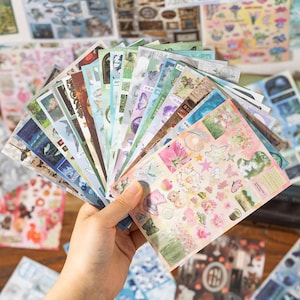 May include: A hand holding a fan of decorative stickers. The stickers feature various designs, including floral patterns, butterflies, and scenic landscapes. The color palette includes pastels, blues, greens, and pinks, creating a whimsical and artistic aesthetic.