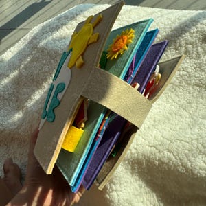 May include: A handmade felt quiet book with a beach theme. The cover features a yellow sun and turquoise waves. Inside, various colorful pages with interactive elements like a lock and other shapes. The book is held together with a beige strap.
