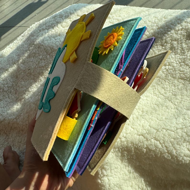 May include: A handmade felt quiet book with a beach theme. The cover features a yellow sun and turquoise waves. Inside, various colorful pages with interactive elements like a lock and other shapes. The book is held together with a beige strap.
