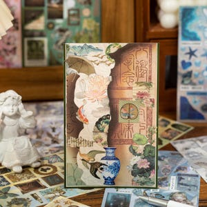 May include: A decorative card with a collage of Asian-inspired imagery. The design includes a blue and white vase, koi fish, lotus flowers, and traditional calligraphy. The card has a green border and a torn paper effect.