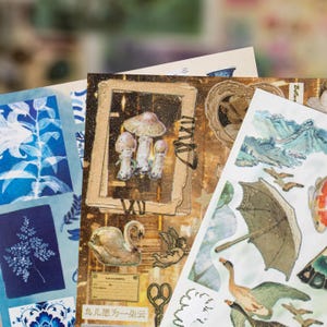 May include: A collection of decorative paper stickers with various designs. The stickers feature illustrations of mushrooms, a swan, an angel, an umbrella, and koi fish. The color palette includes shades of blue, brown, and gold, with some stickers having a vintage aesthetic.