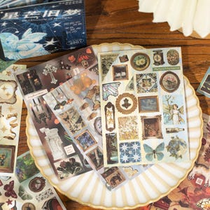 May include: A collection of decorative stickers with vintage-style illustrations, including framed art, butterflies, and floral designs. The stickers are arranged on a decorative plate, with a box of stickers in the background. The color palette includes beige, brown, and blue.