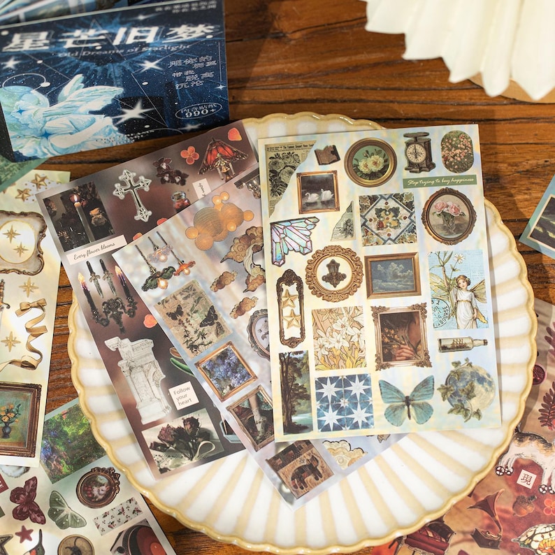 May include: A collection of decorative stickers with vintage-style illustrations, including framed art, butterflies, and floral designs. The stickers are arranged on a decorative plate, with a box of stickers in the background. The color palette includes beige, brown, and blue.