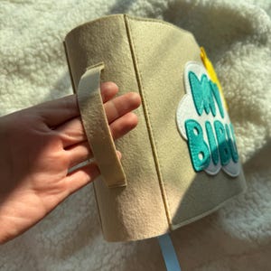 May include: A beige felt book with the words "MY BIBU" in turquoise and white, with a yellow accent. The book has a handle and a light blue ribbon bookmark.